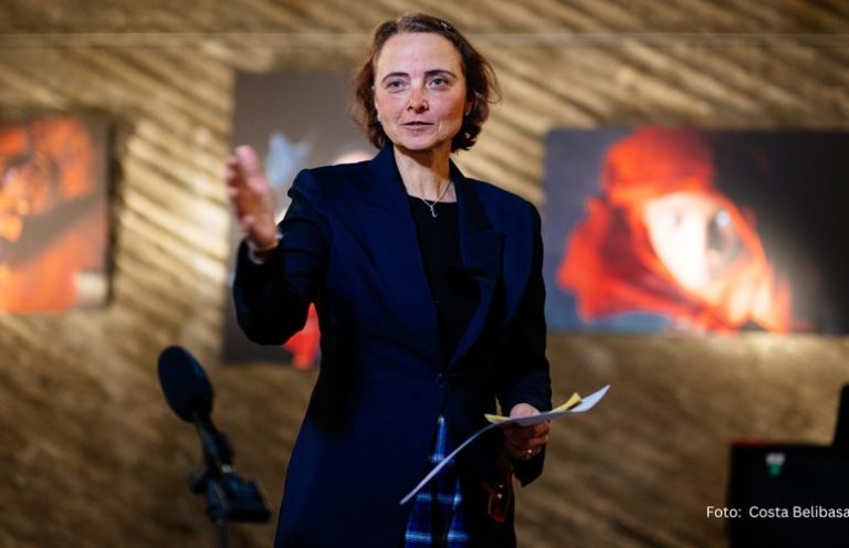 Die Kuratorin Seona Sommer spricht mit gestrecktem Arm, im Hintergrund hängen Kunstwerke.
