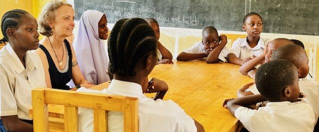 Die Künstlerin und Aktivistin Seona Sommer umgeben von Schülern in einem Klassenzimmer in Tansania, alle hören aufmerksam zu.